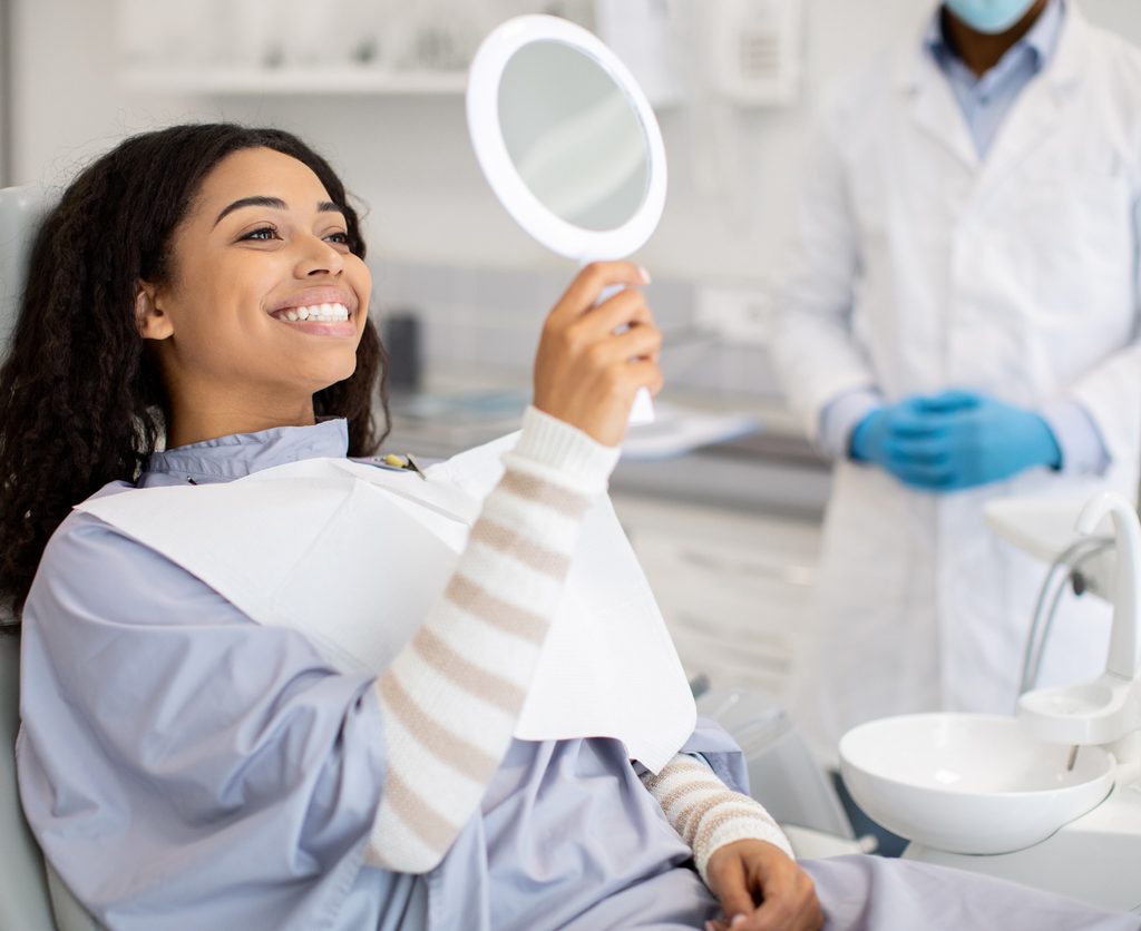 Happy Female Patient Looking At Mirror After Dental Treatment In Clinic